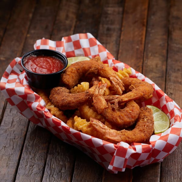 Fried shrimp and waffle fries in a red and white checkered basket with ketchup and lime.