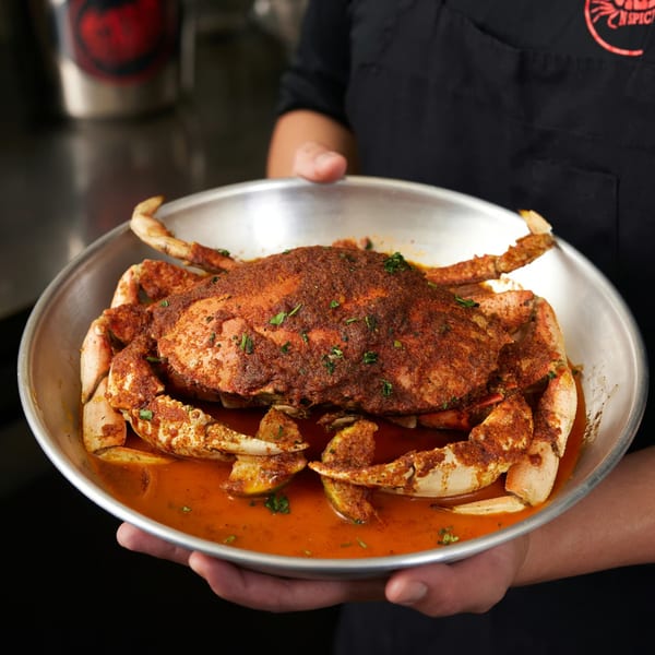 A person holding a large metal bowl filled with a whole seasoned crab in a rich red sauce.