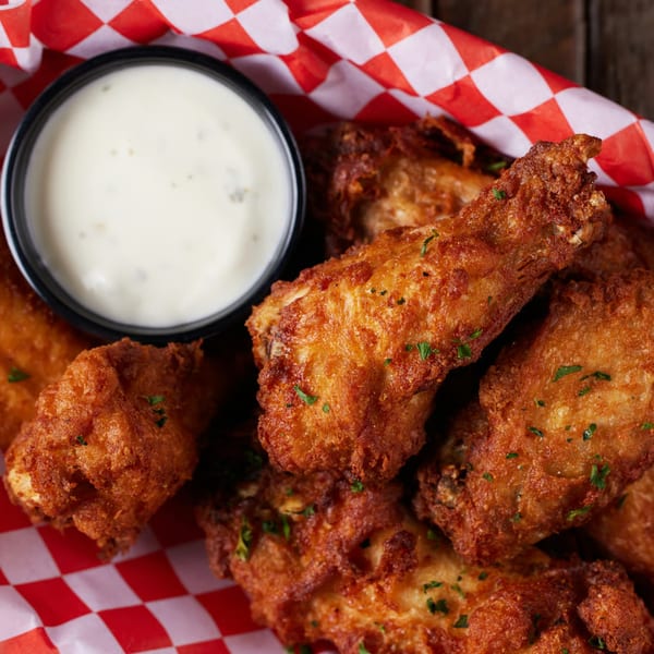 Crispy fried chicken wings with parsley flakes and a side of creamy ranch dressing in a red checkered basket.